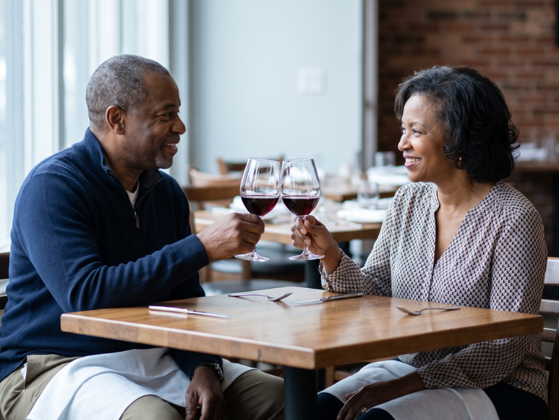 Smiling couple sitting at a restaurant table, toasting with glasses of red wine.