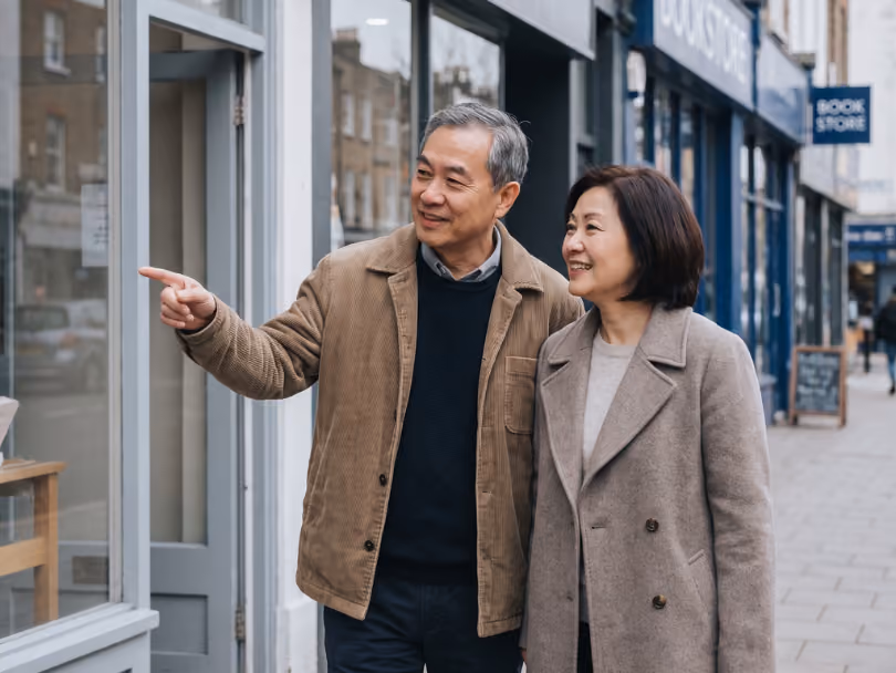 Smiling middle-aged couple looking and pointing at a shop window on a city sidewalk.