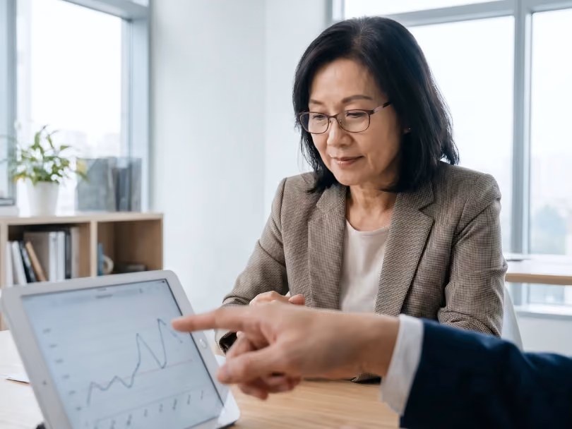 Businesswoman in glasses listens as colleague points to a line graph on a tablet during meeting.
