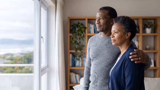 Middle-aged couple standing by a window, looking outside with a calm expression in a cozy room.