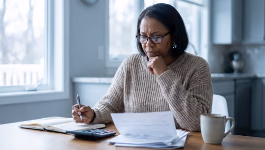 Woman wearing glasses reviewing documents with a pen, calculator, and coffee cup at a table.