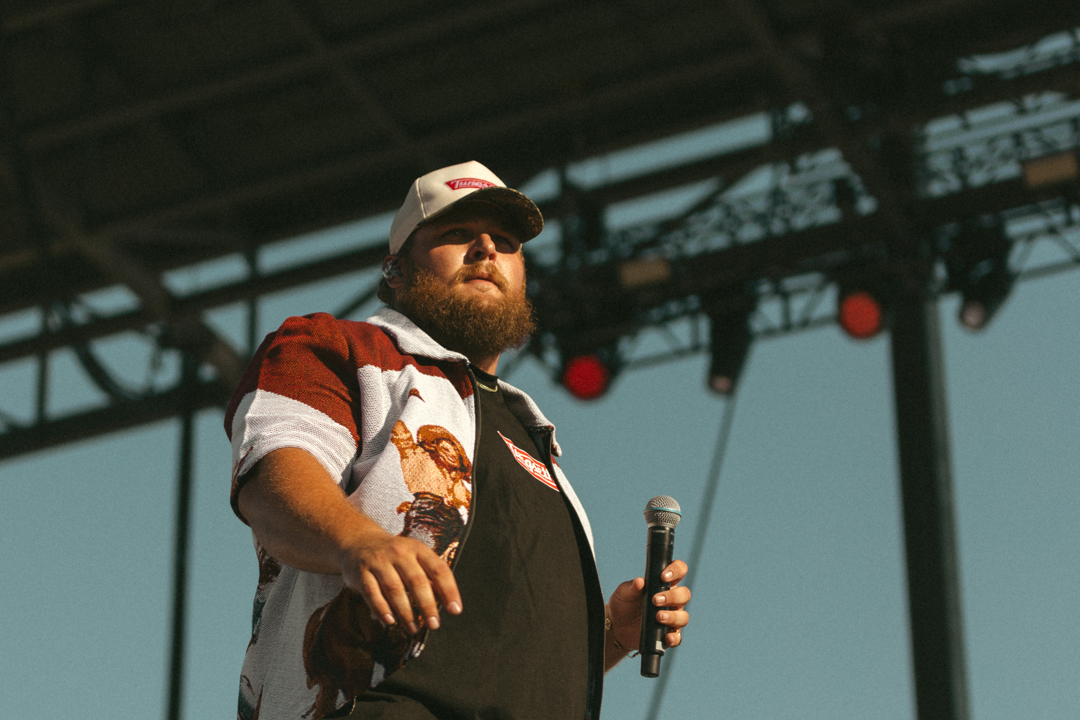 Chris Tungseth wearing a white and red patterned short-sleeve shirt and a white cap holding a microphone on stage under a metal framework with red stage lights.