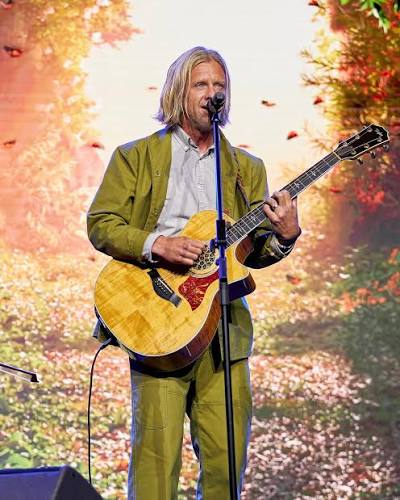 Jon Foreman shown with shoulder-length blonde hair wearing a green suit playing an acoustic guitar and singing into a microphone on stage with a backdrop of autumn foliage.