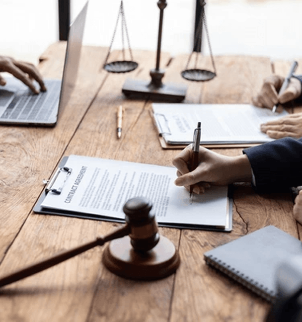 Hands signing a contract on a clipboard with a gavel and scales of justice on a wooden table.