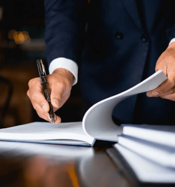 Person in suit signing or reviewing documents on a desk with a pen in hand.
