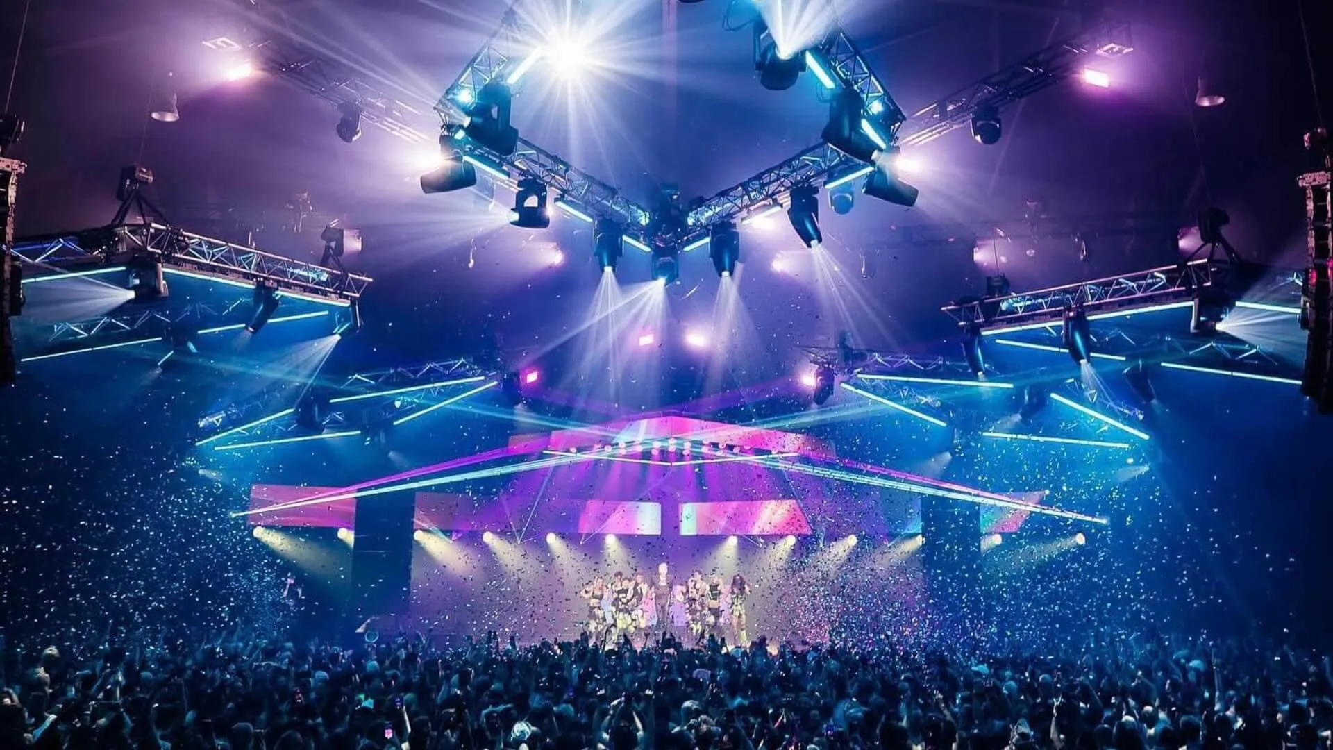 Large crowd surrounds a circular concert stage beneath colourful lights and lasers.