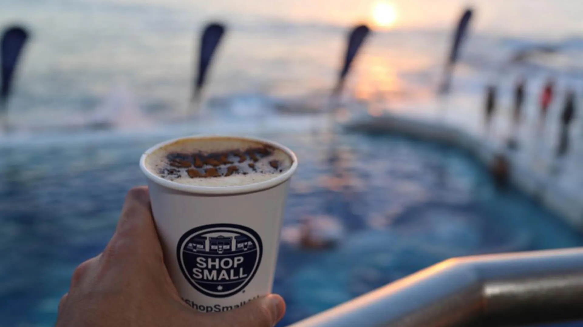 Hand holds a Shop Small branded coffee cup beside the ocean pool.