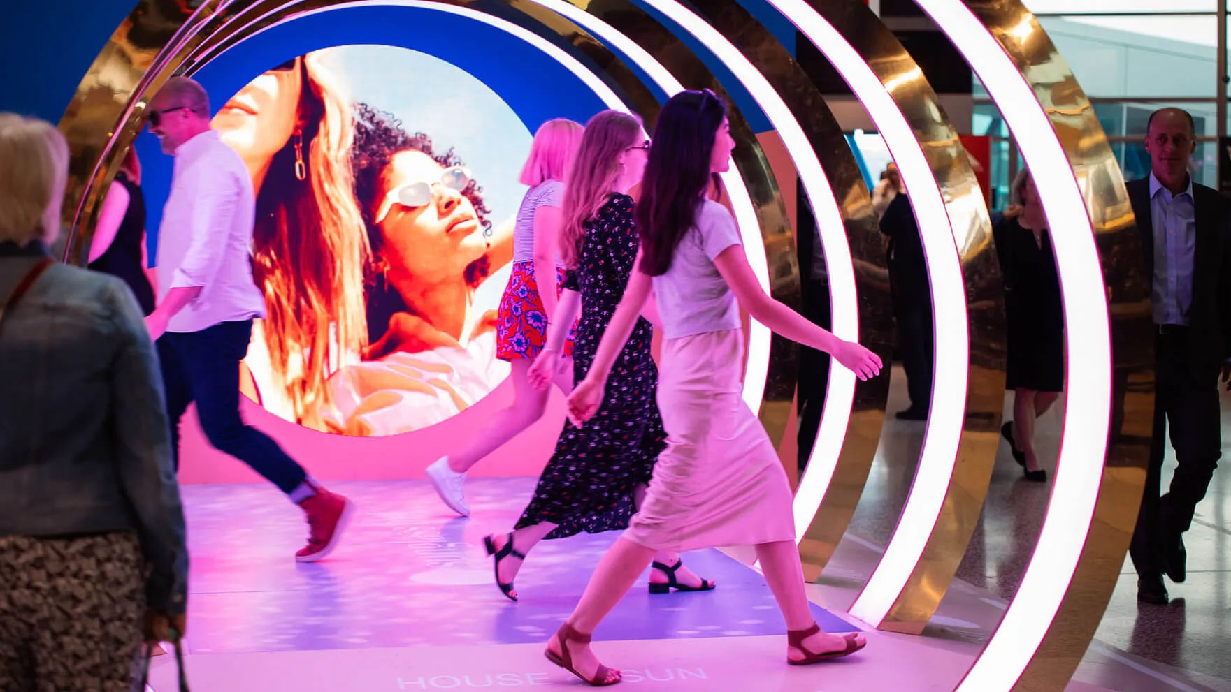 Visitors walk through illuminated arches with a large portrait display behind them.