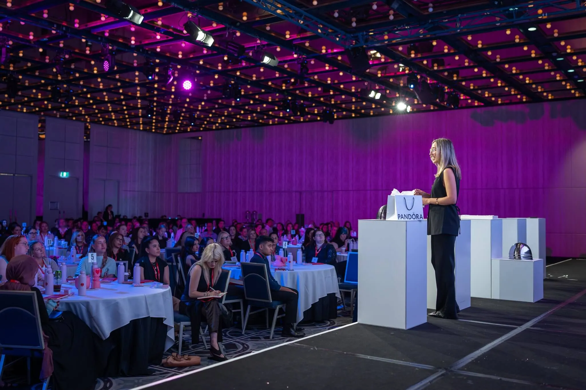 Presenter addresses seated delegates from a white podium at the Pandora conference.