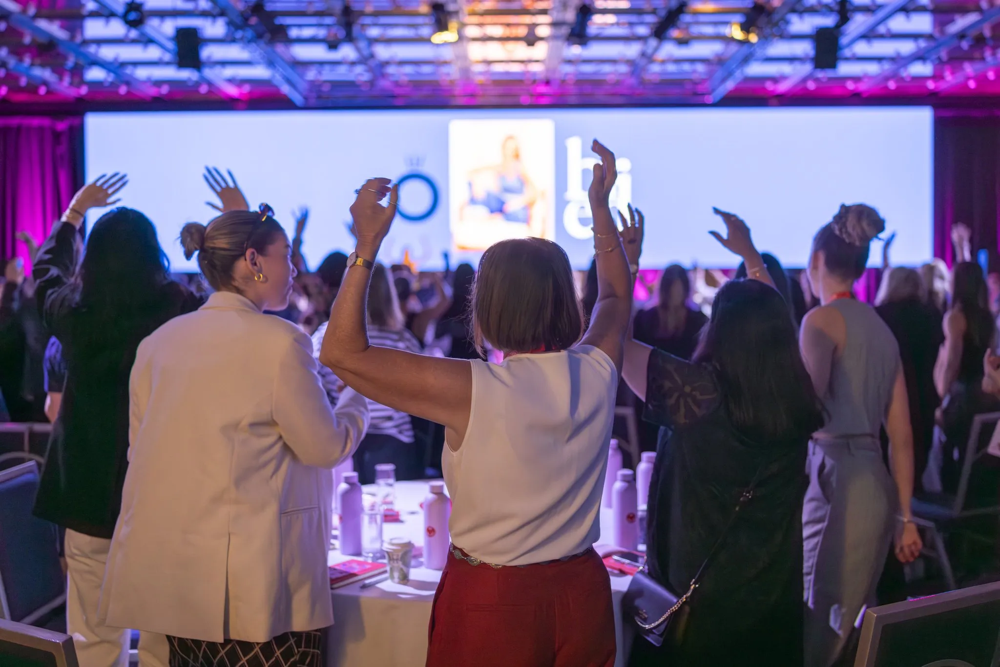 Conference attendees stand and raise their hands beneath the illuminated stage screen.