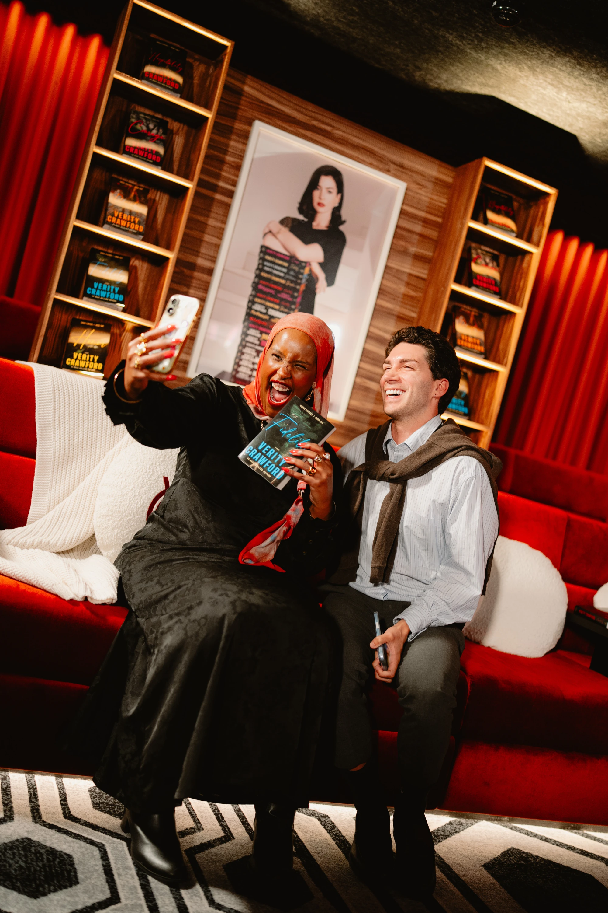 Two guests pose together in a lounge with bookshelves and red curtains at the Verity event.