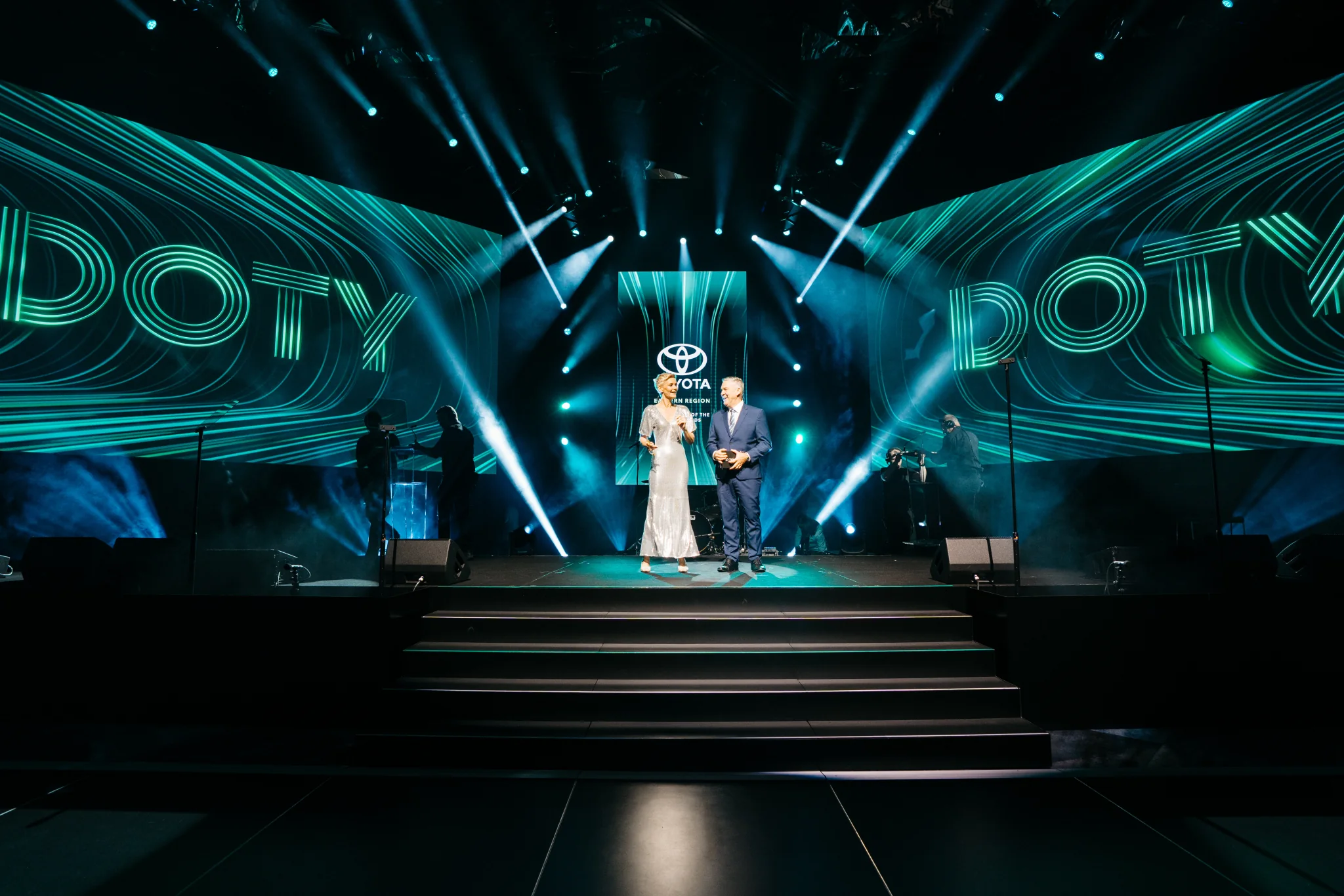 Two hosts on the Toyota Dealer of the Year stage beneath green event screens.