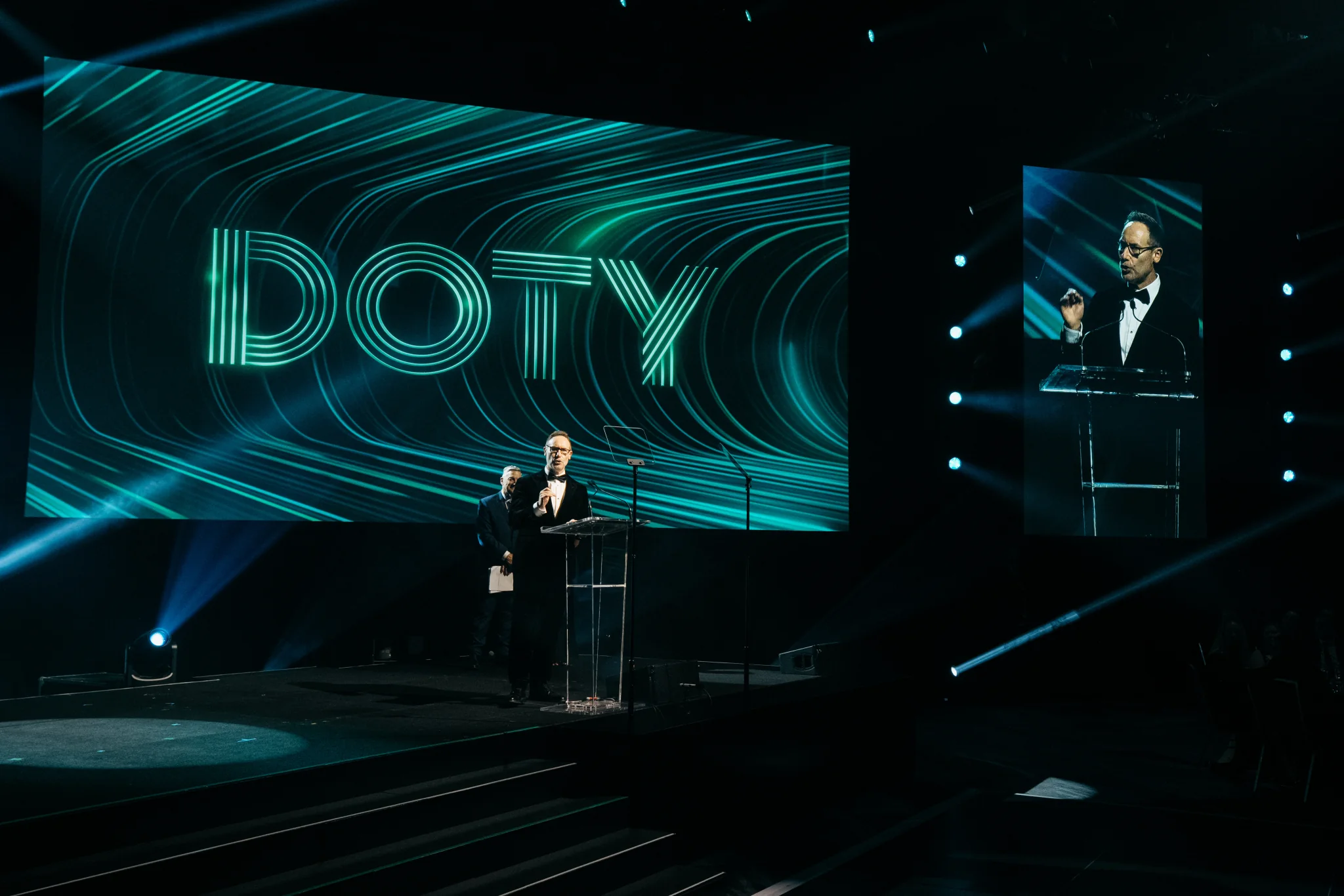 A speaker at a clear lectern with a live-feed portrait on the Toyota awards stage.