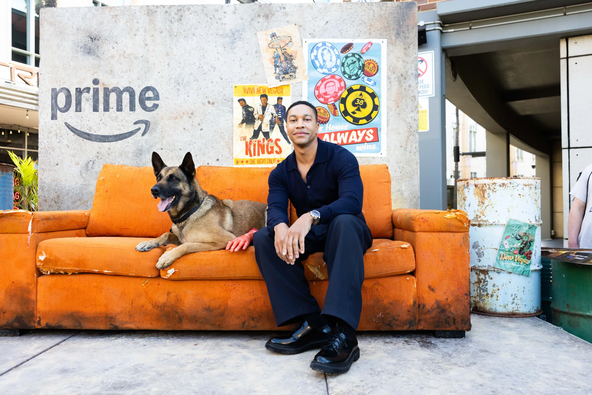 A cast member sits with a German shepherd on an orange sofa in the outdoor Fallout set.