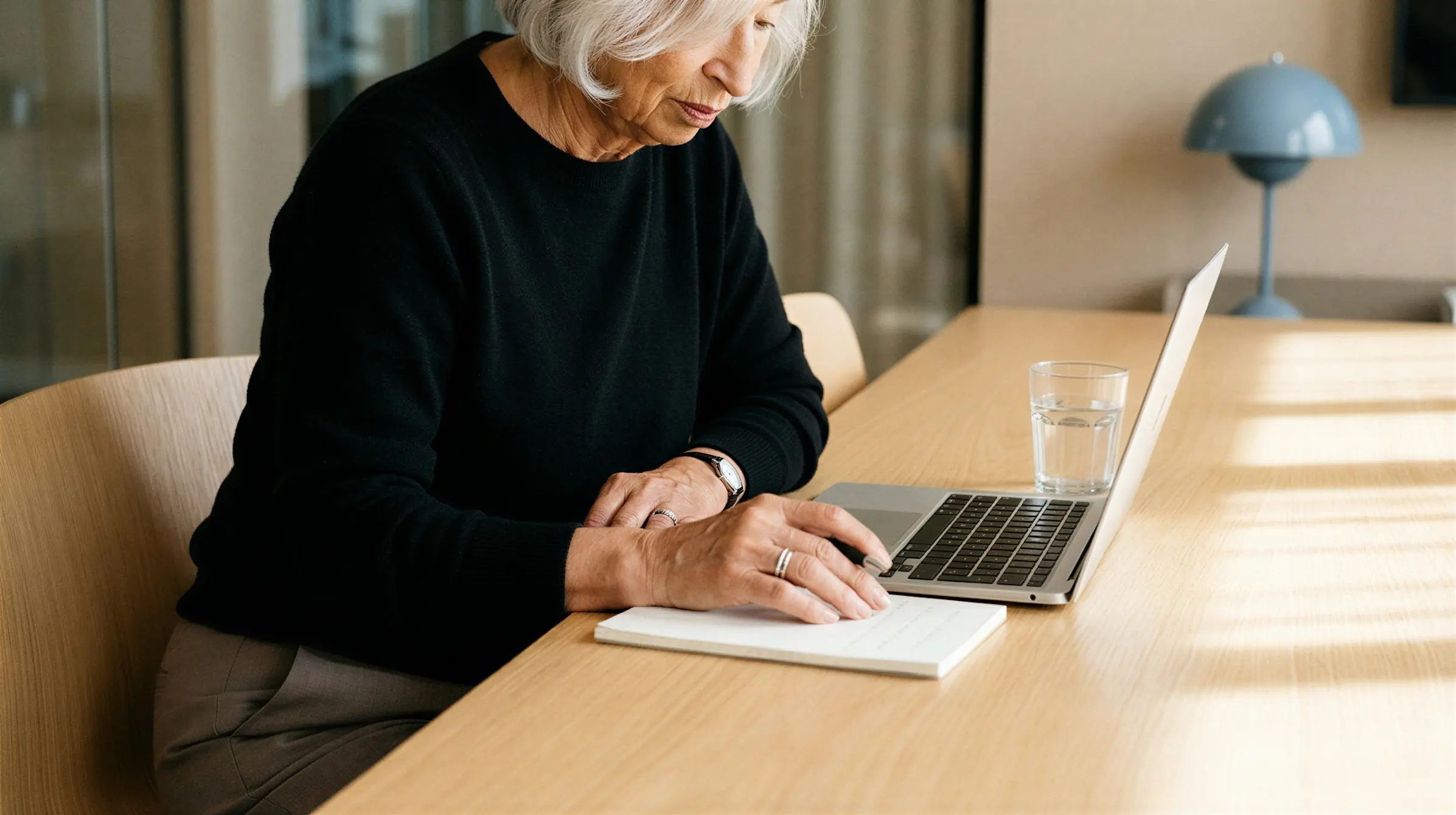 Elderly woman in a black sweater reading notes near a laptop with a glass of water on a wooden table