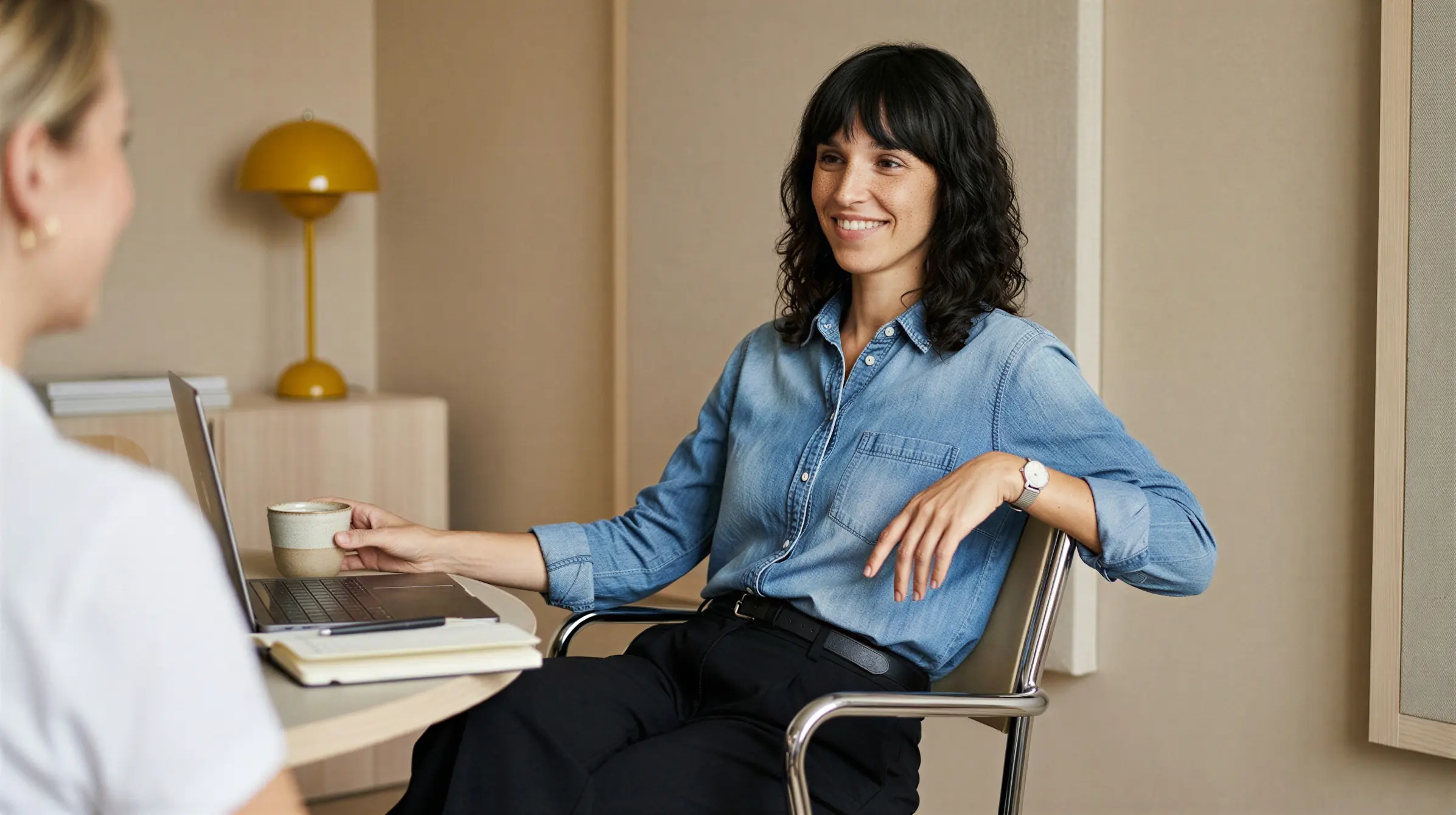 Woman smiling in a denim shirt sitting in a chair with a laptop, coffee cup, and notebook on the table