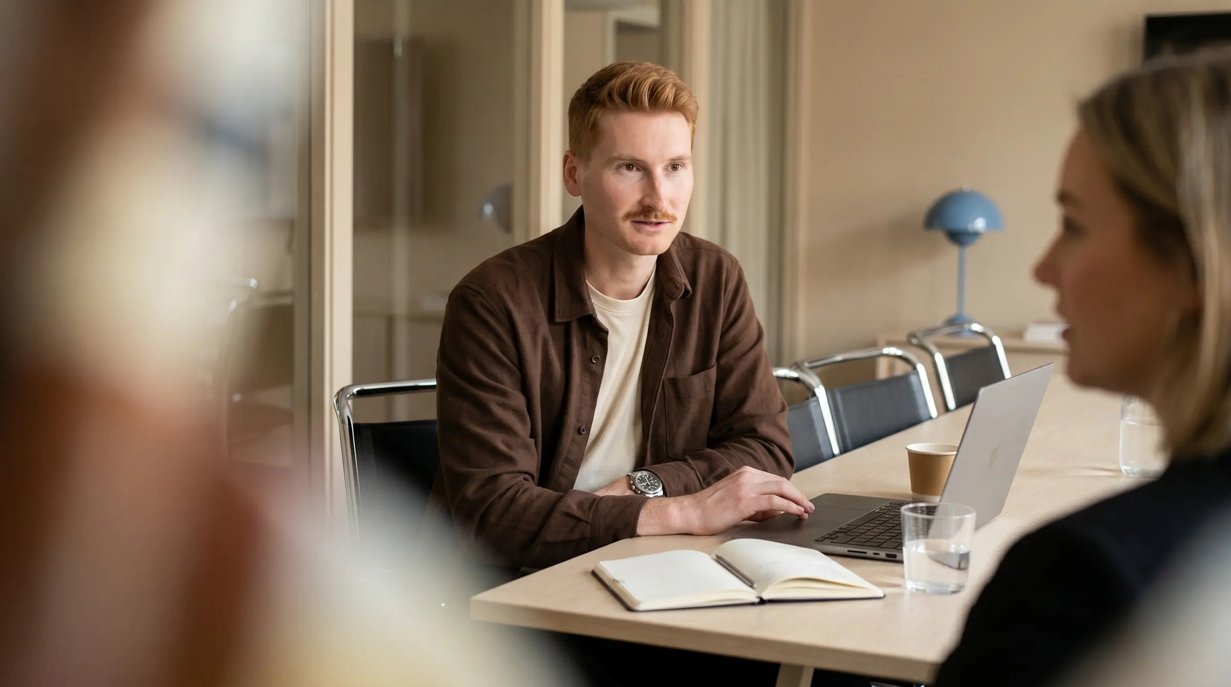 Man with red hair and a mustache talking to two colleagues at a conference table with a laptop and notebook