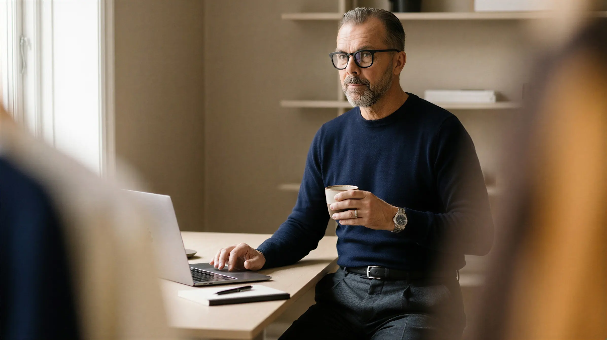 Man in glasses sitting at a desk using a laptop and holding a cup of coffee