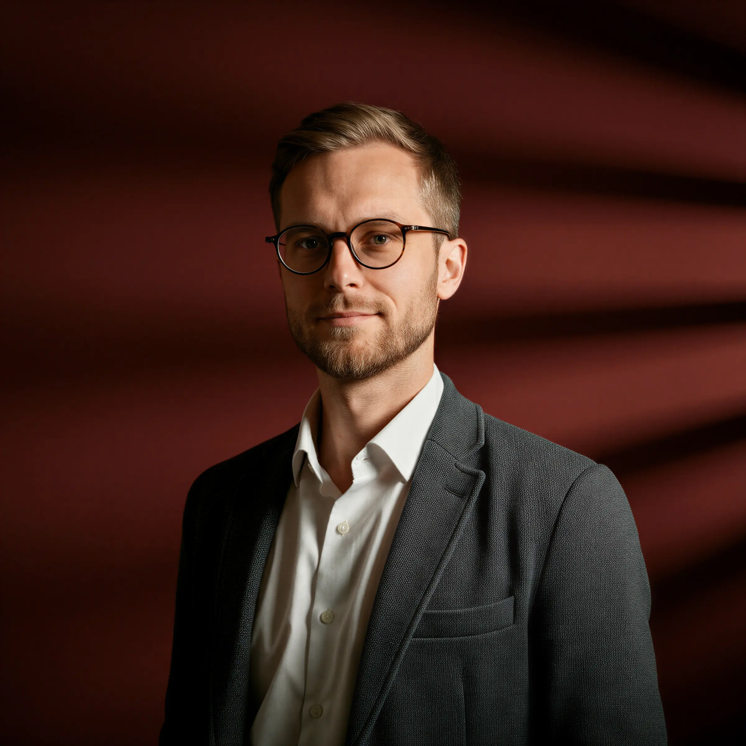 Portrait of a man with glasses in a gray blazer and white shirt against a dark red background
