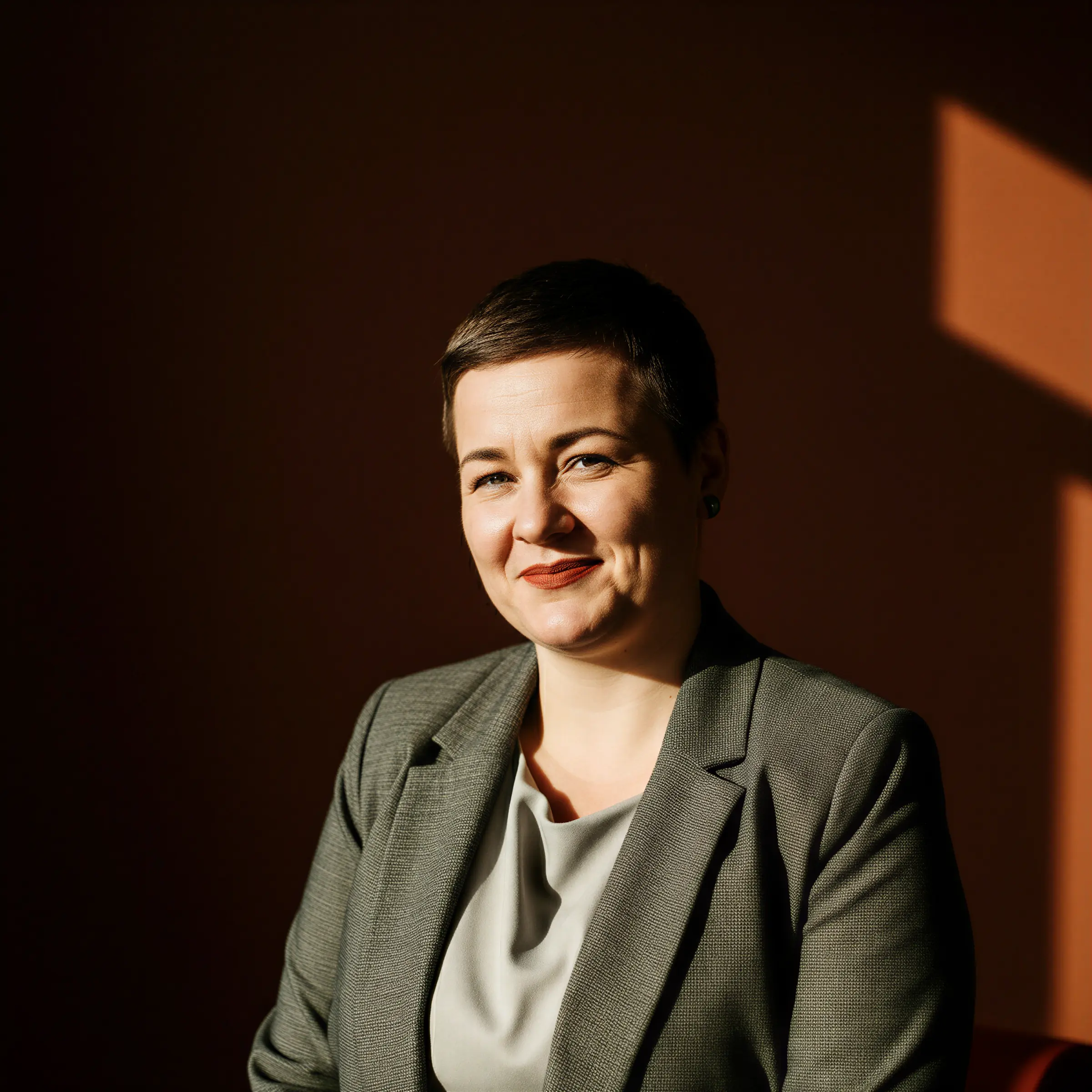 Smiling woman with short hair wearing a gray blazer and light gray blouse against a dark background