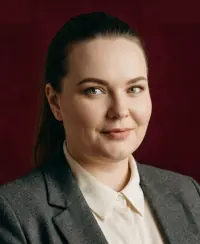 Smiling woman with green eyes and dark hair in a gray blazer and white shirt against a maroon background.