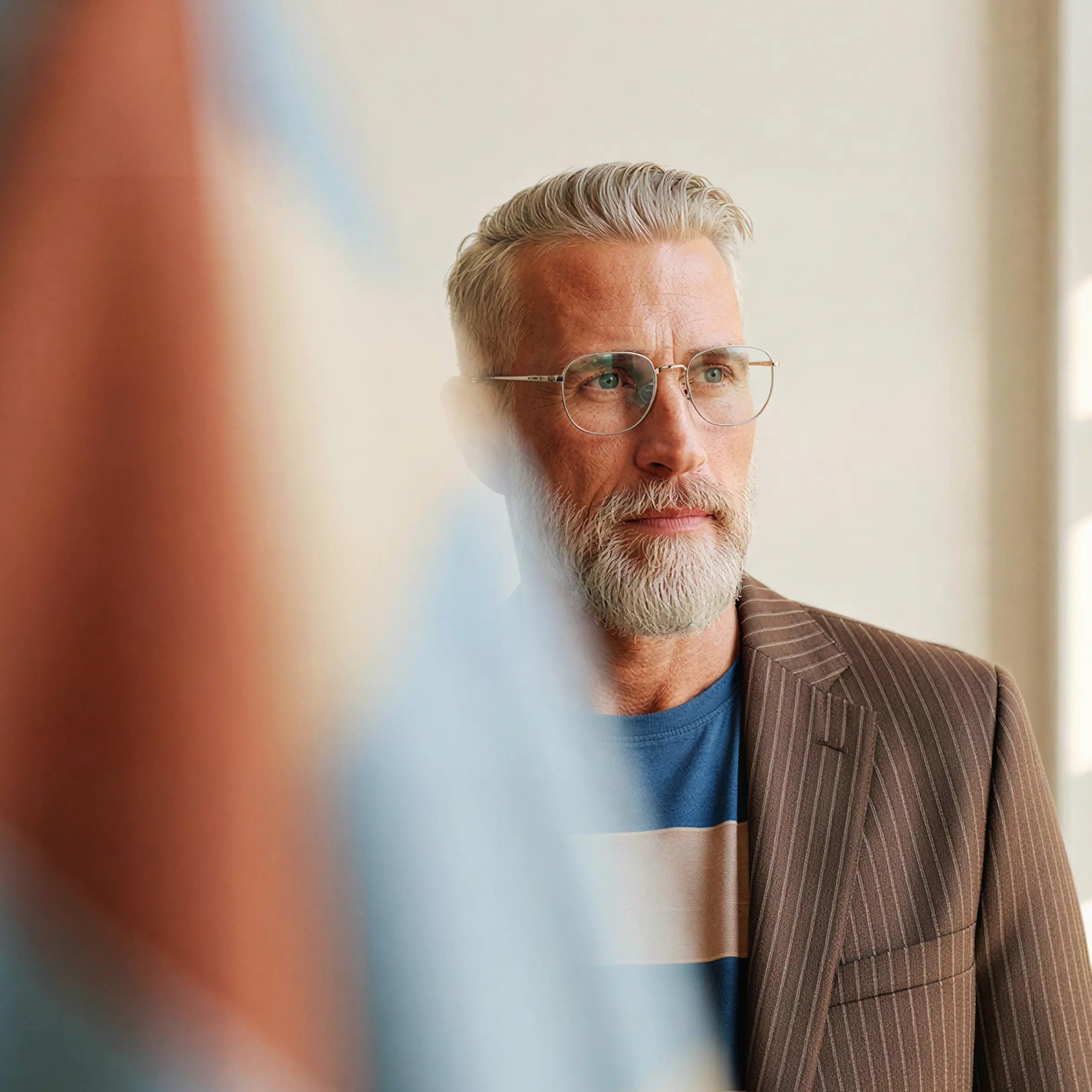 Contemplative gray-haired man with beard and glasses wearing a striped brown jacket and blue shirt.