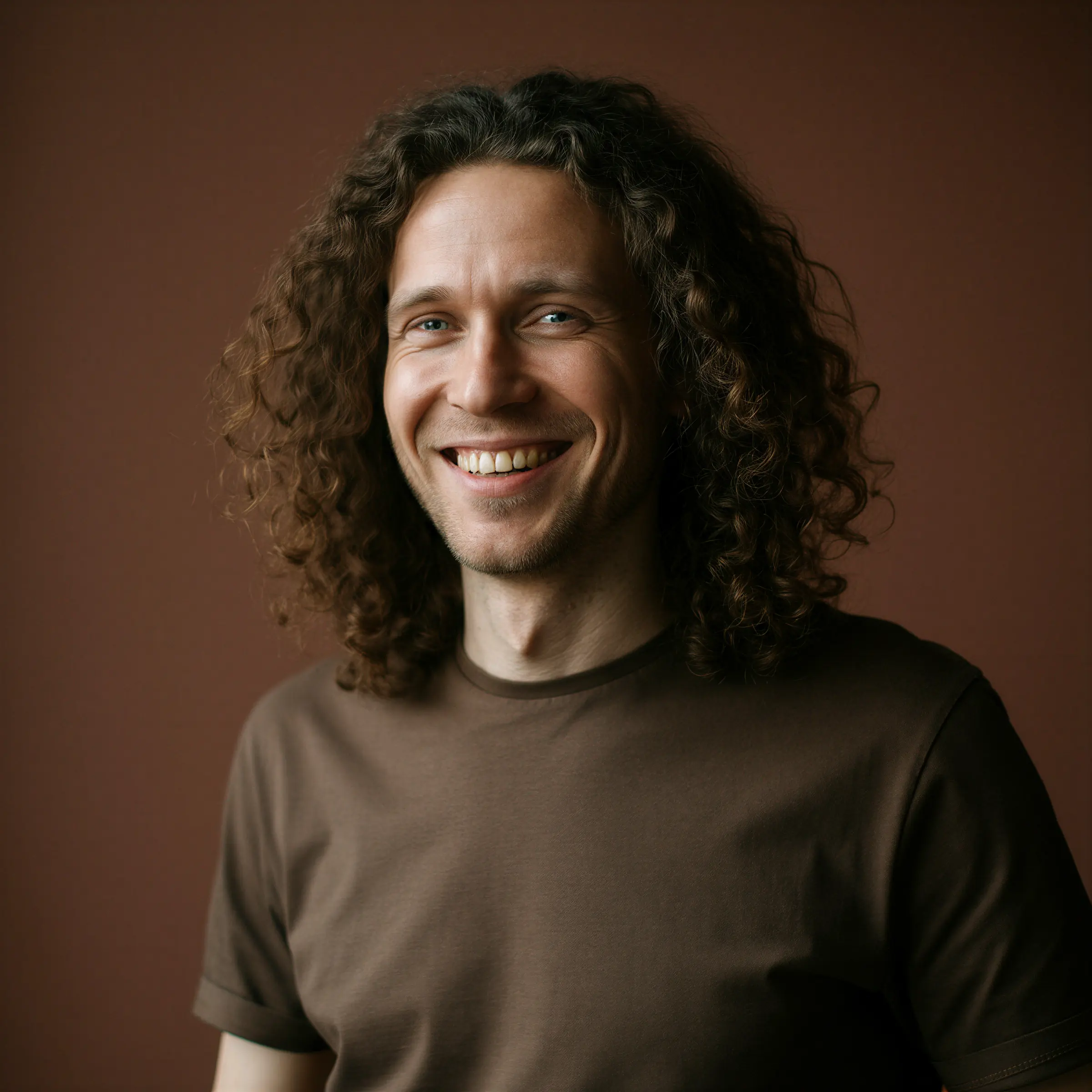Smiling man with long curly hair wearing a brown t-shirt against a brown background.