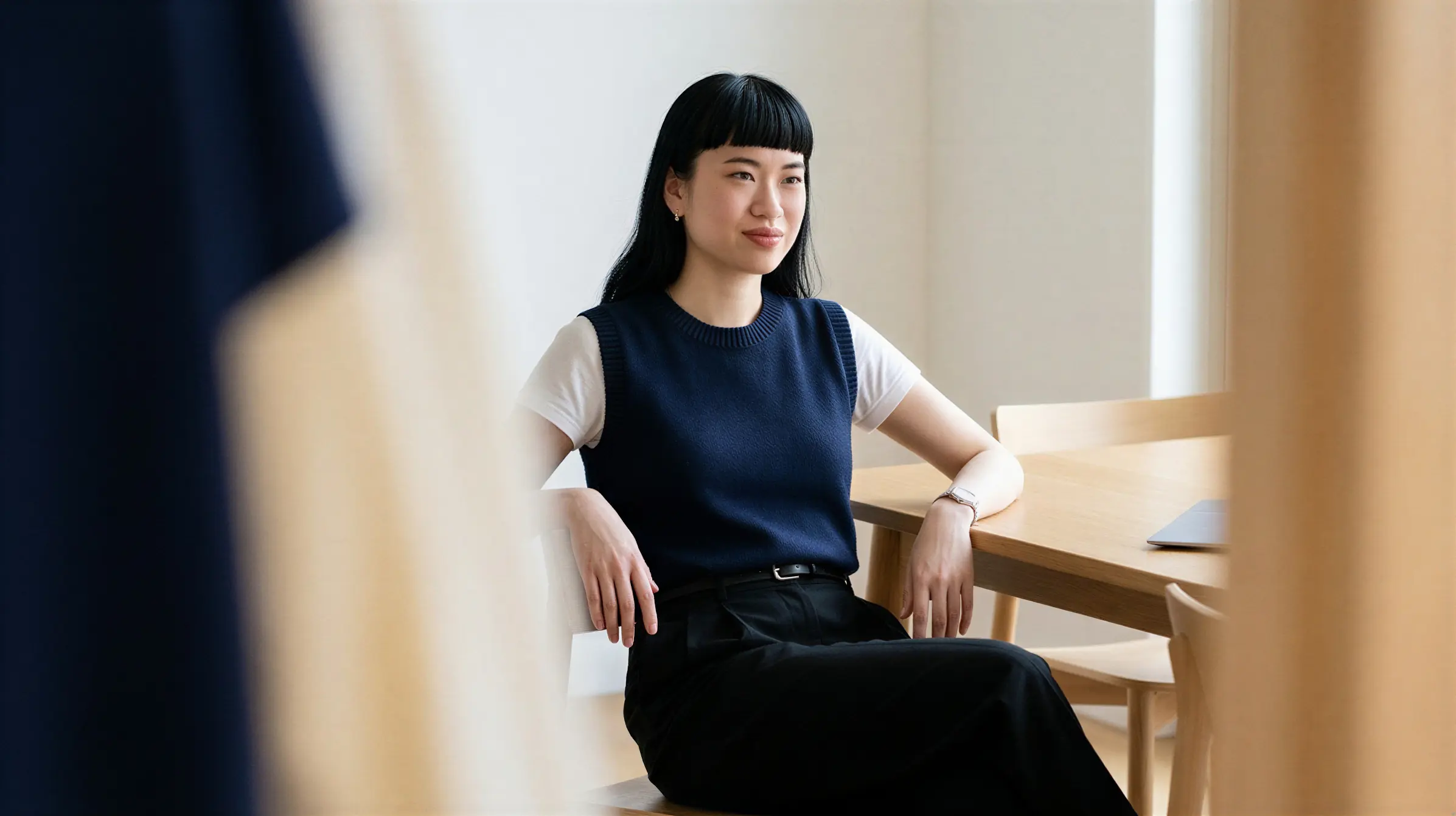 Young woman with black hair sitting at a wooden table looking thoughtfully to the side.