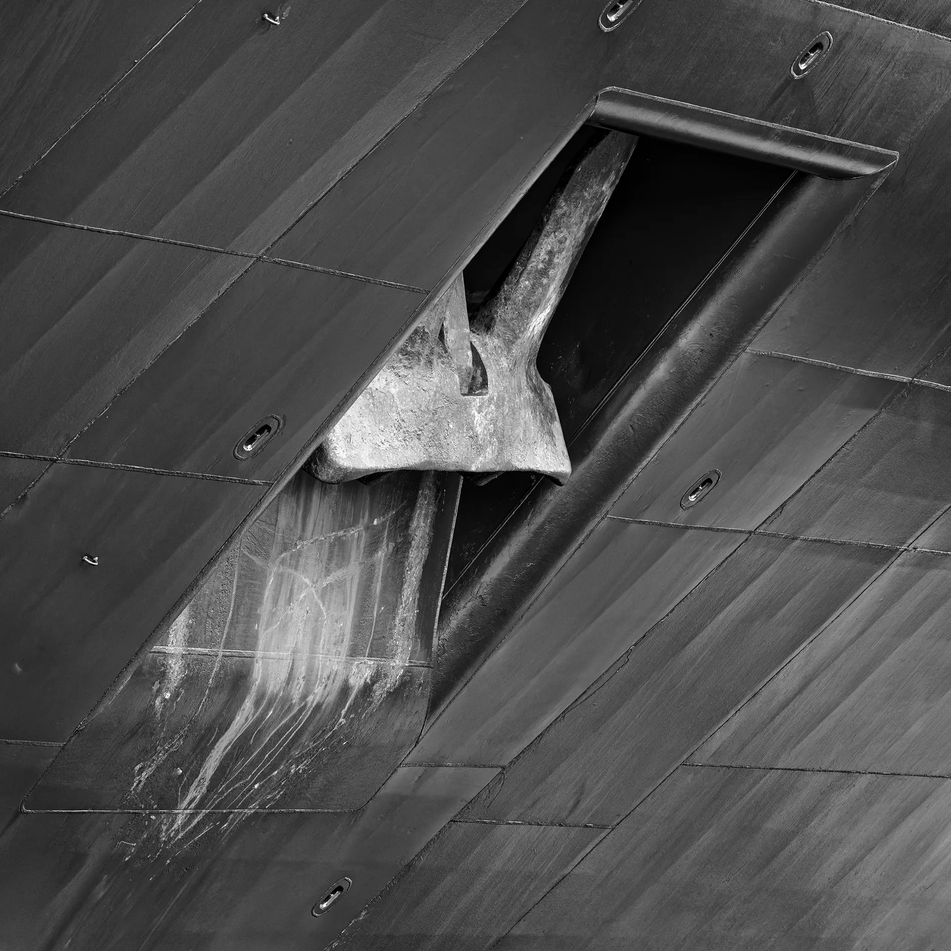 A black-and-white close-up of a ship's anchor stowed in its hawsepipe against the steel hull plating, with staining streaked down below it.