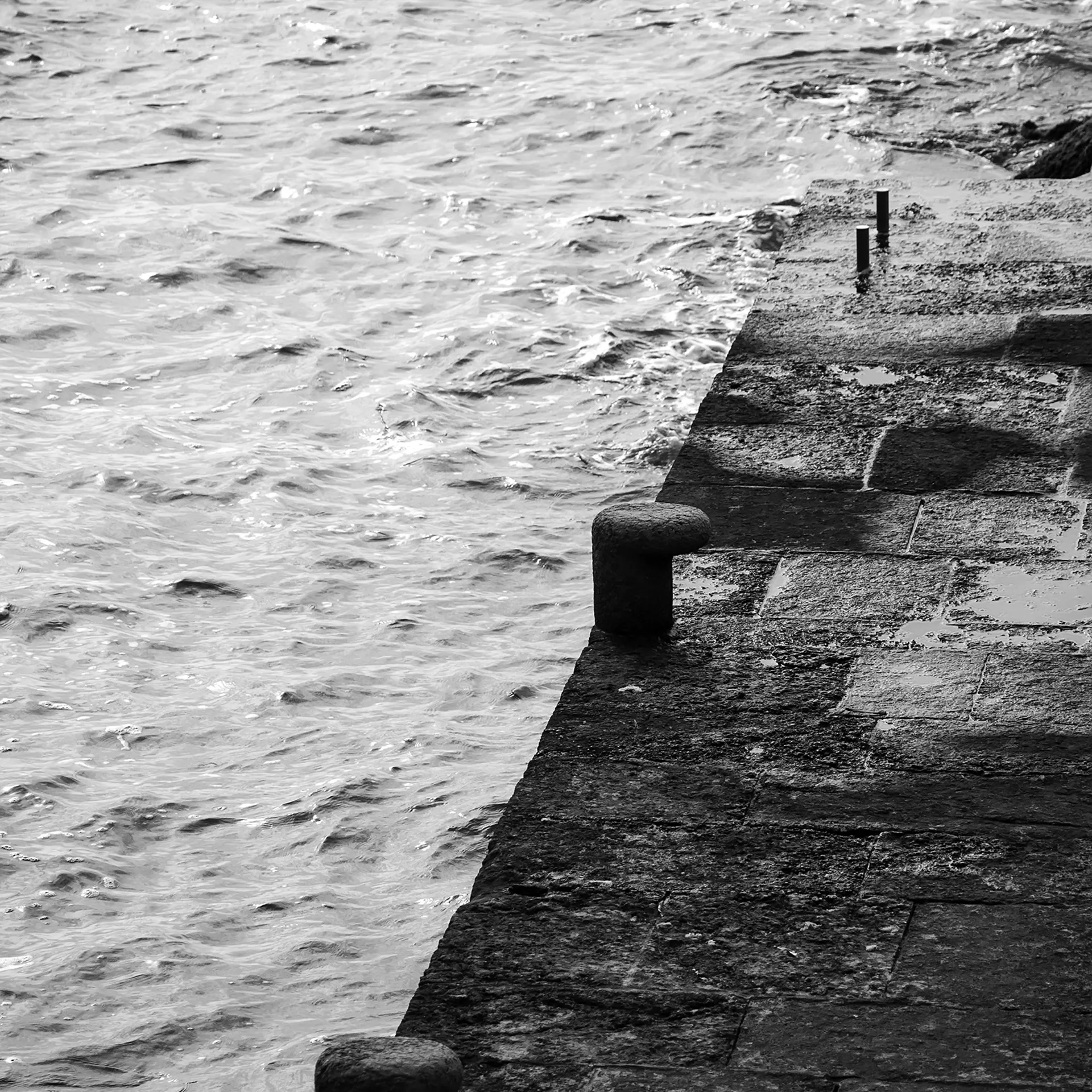 A stone pier with mooring bollards running diagonally alongside choppy gray water, bordered by a rocky embankment.