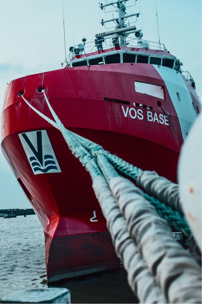 The bow of a red and white offshore supply vessel named "VOS BASE" moored at a dock, with thick mooring ropes running from the ship to the quay in the foreground.