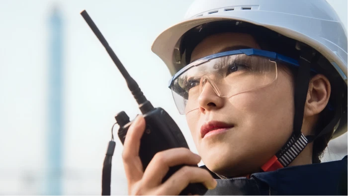 A worker wearing a white hard hat and safety glasses holding a two-way radio, with an industrial structure blurred in the background.