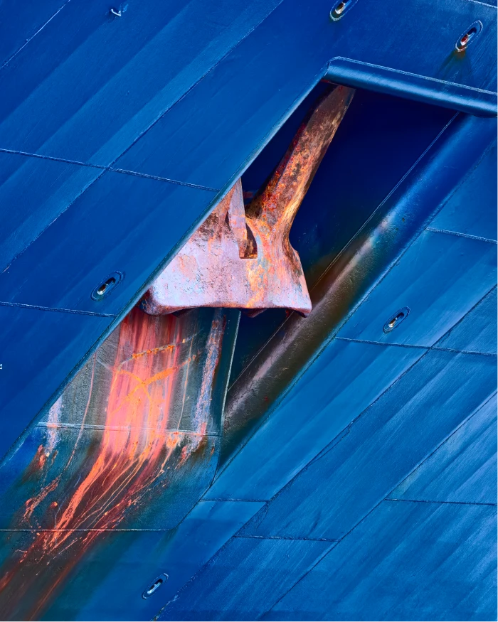 A close-up of a ship's anchor stowed in its hawsepipe against a blue steel hull, with orange rust streaks running down the plating.