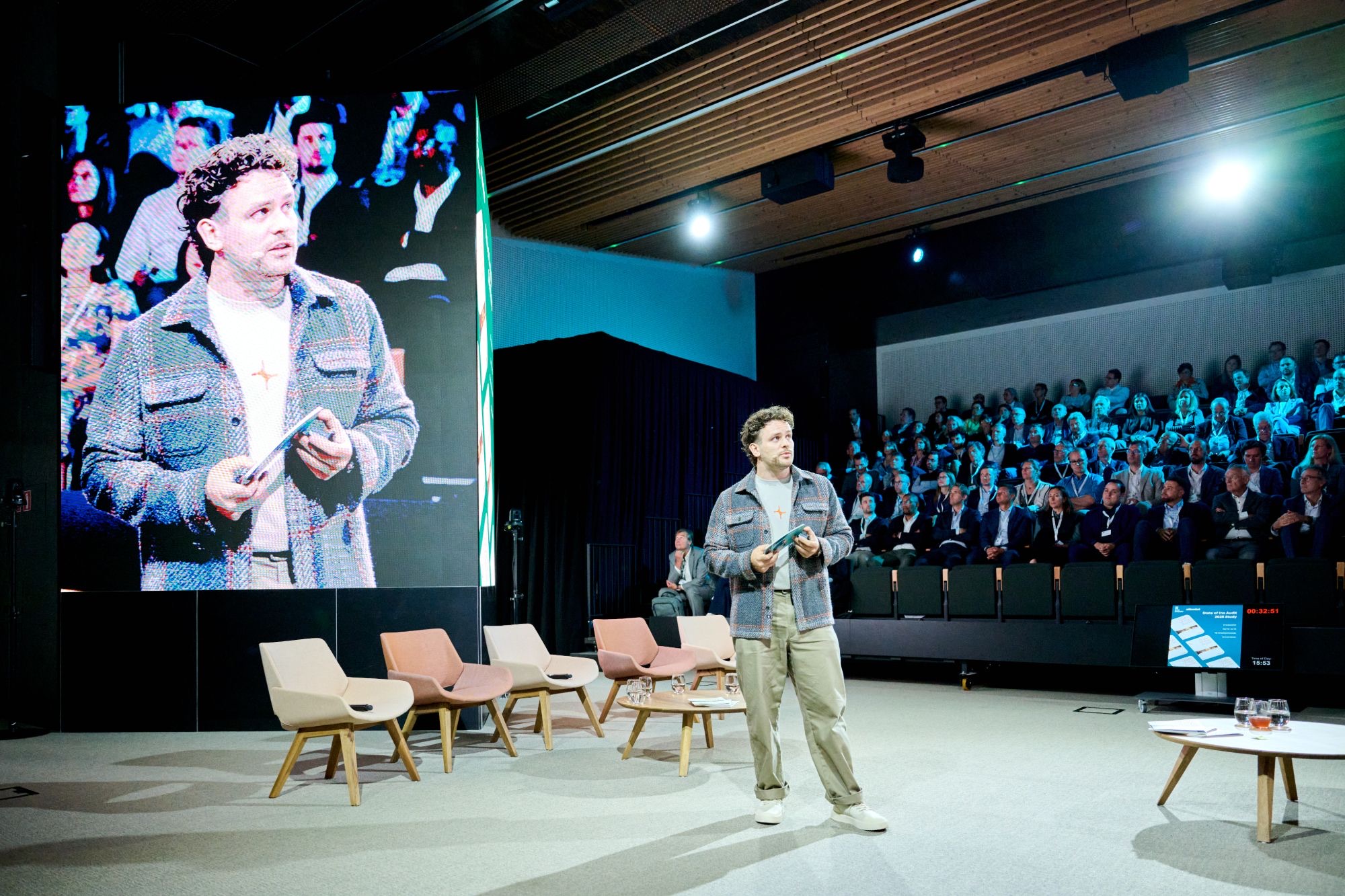 dr. Mathias Celis on stage in front of a full auditorium, his image on the large screen behind him