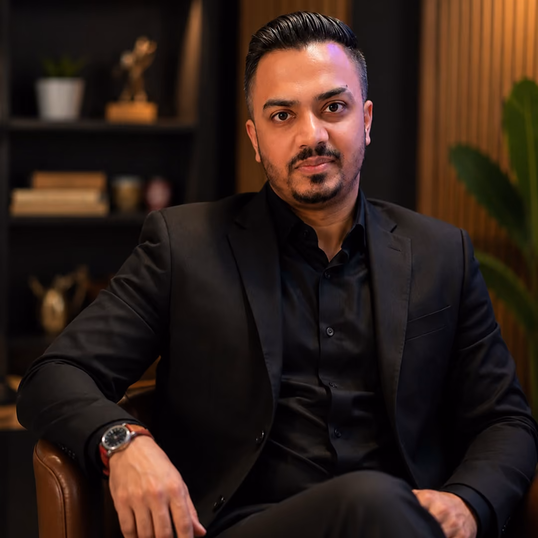 Man in a black suit sitting in a brown chair with blurred indoor background.