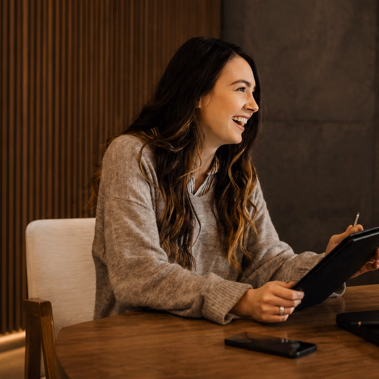 Smiling woman with long hair in a beige sweater holding a tablet and stylus at a wooden table.