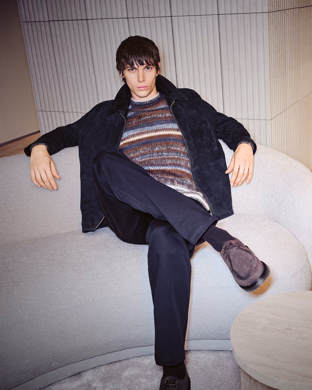 Model in gestreiftem Strickpullover und dunkler Jacke auf einem hellen Sessel
