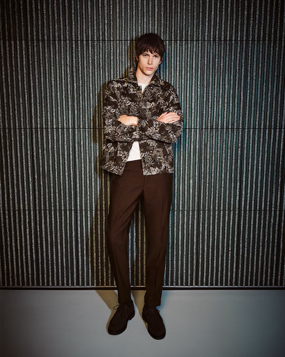 Model in gemusterter Hemdjacke und brauner Hose vor einer dunkelgrünen Wand