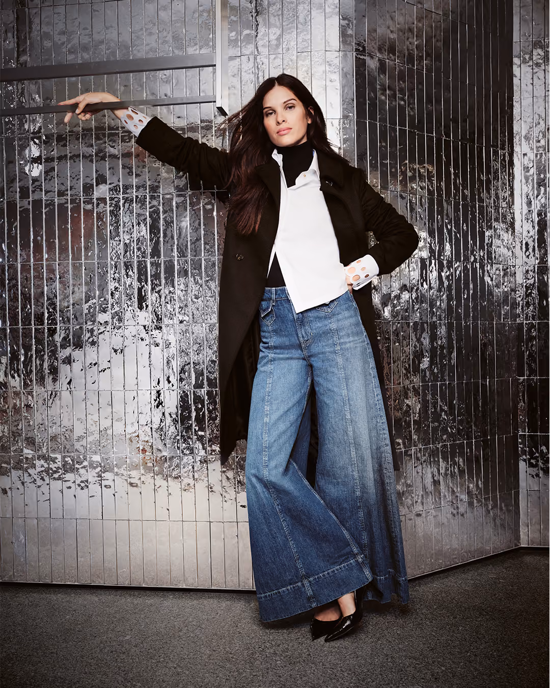 Model in schwarzem Mantel und weiter Jeans vor einer silbernen Wand