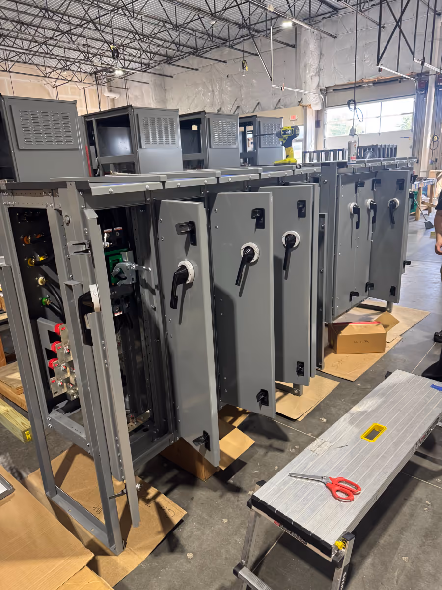 Row of grey industrial electrical panels with open doors in a warehouse with tools nearby.