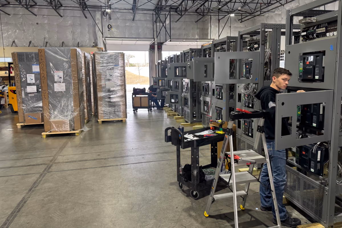 Worker in a warehouse assembling or inspecting large electrical control panels lined up in rows.
