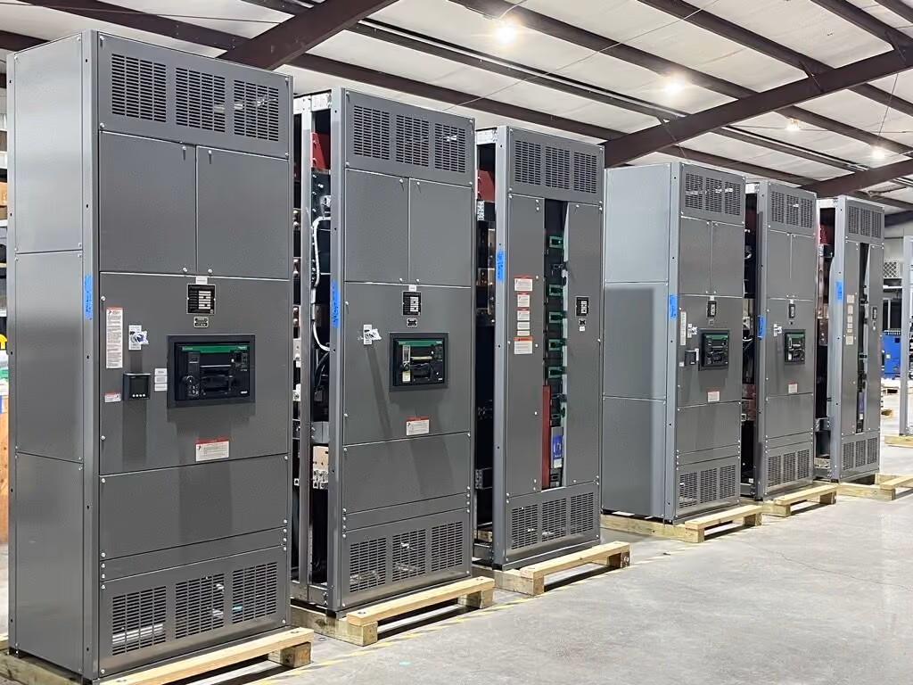 large grey industrial electrical panels side by side, each with multiple black circuit breaker switches and vents, placed on wooden pallets inside a warehouse with metal ceiling beams visible.