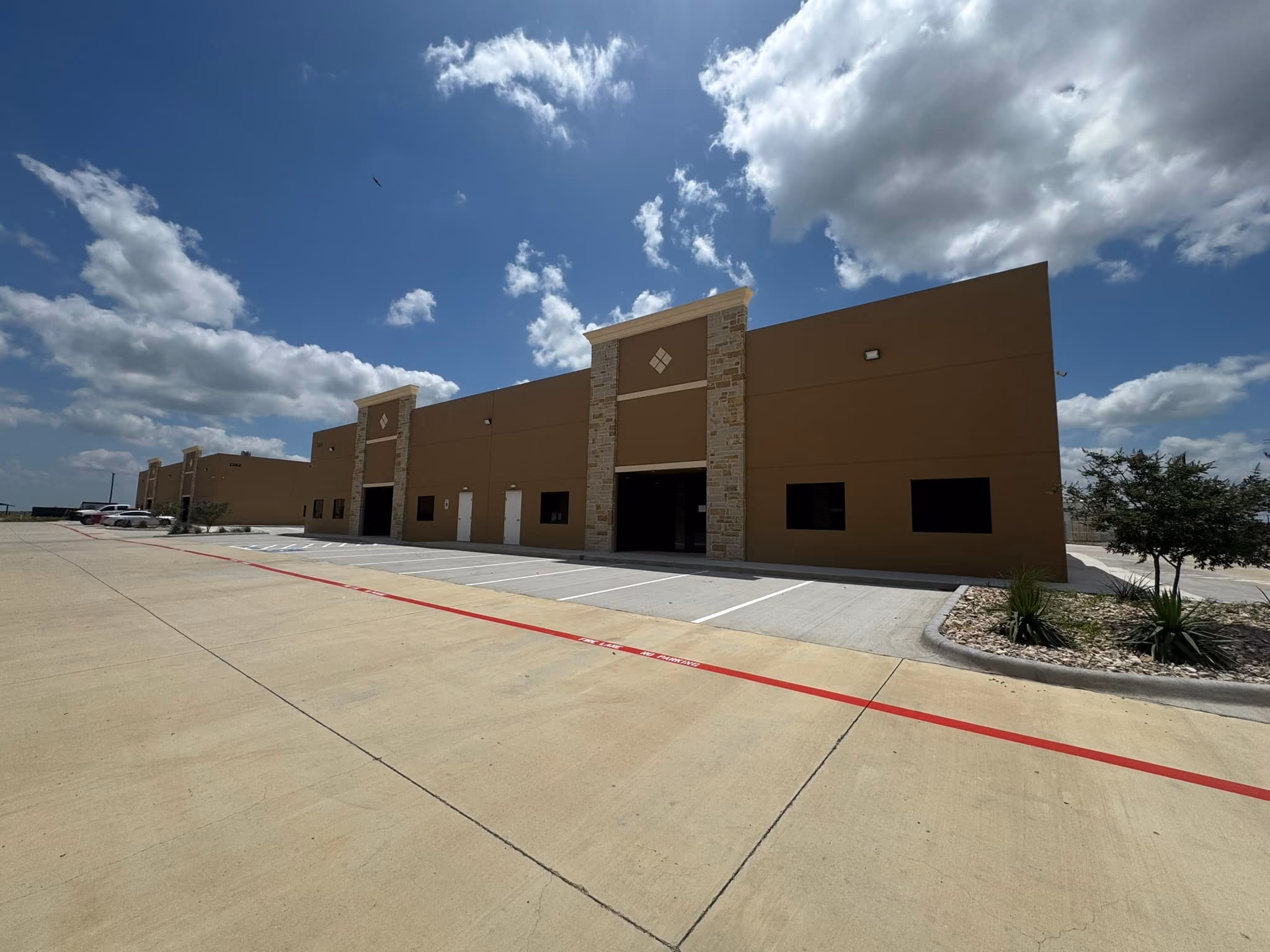 Long brown commercial warehouse building with stone accents under a partly cloudy blue sky, empty parking spaces, and small landscaped areas with plants on the right side.