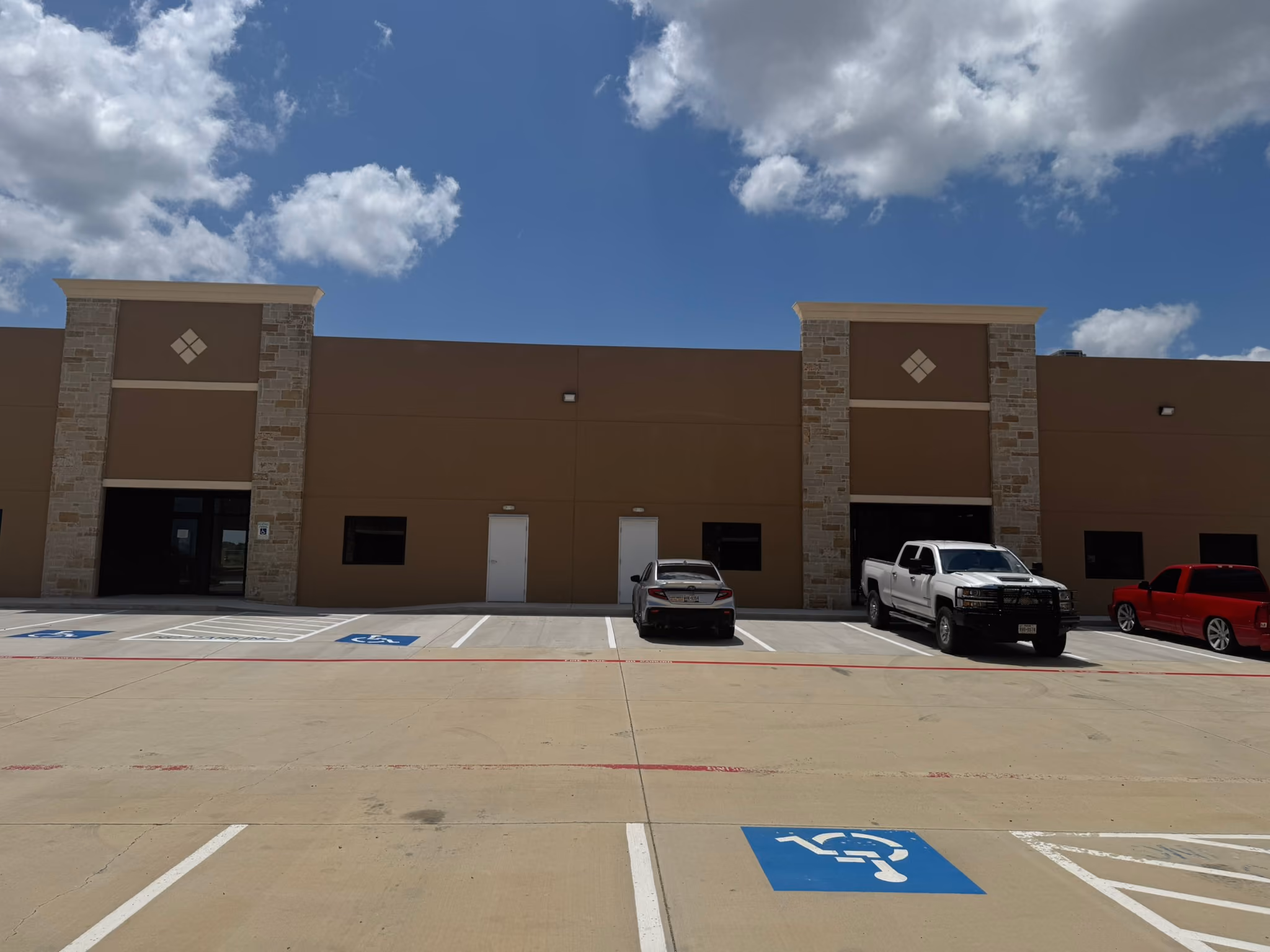 Parking lot in front of a brown commercial building with stone pillars, partly cloudy blue sky, and three vehicles parked, including a gray car, a white truck, and a red vehicle.