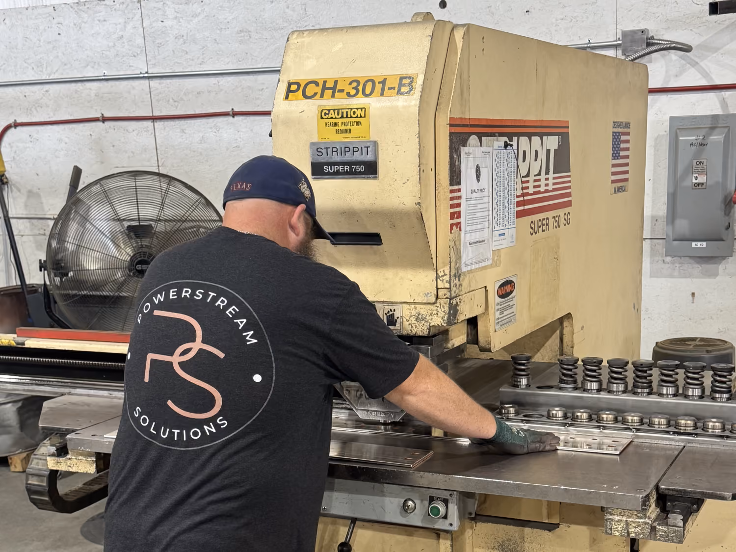 Man operating a large industrial metal cutting machine labeled 'STRIPPIT SUPER 750' in a workshop, wearing a black t-shirt with 'POWERSTREAM SOLUTIONS' logo on the back and a navy blue cap.