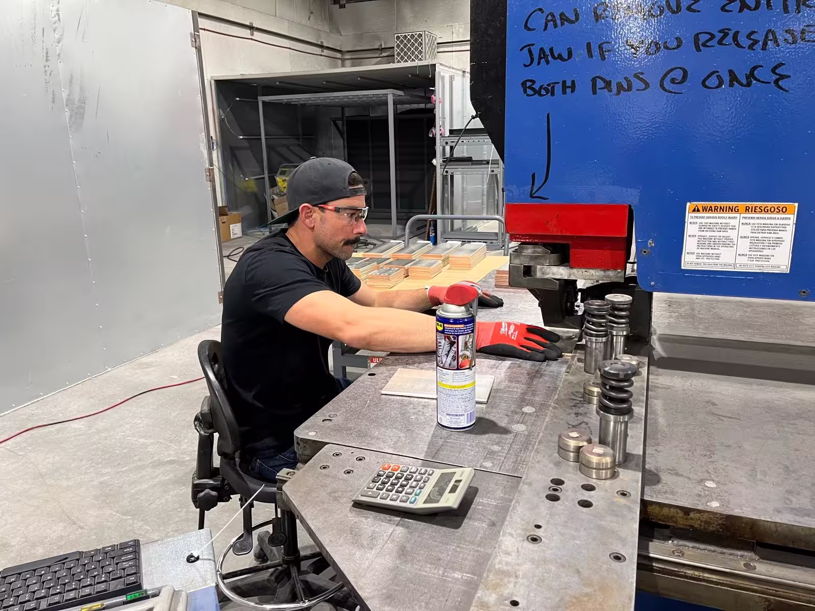 Man wearing safety glasses, a backwards cap, and red gloves operates a blue industrial machine in a workshop, with metal parts, a calculator, and a spray can on the workbench in front of him.