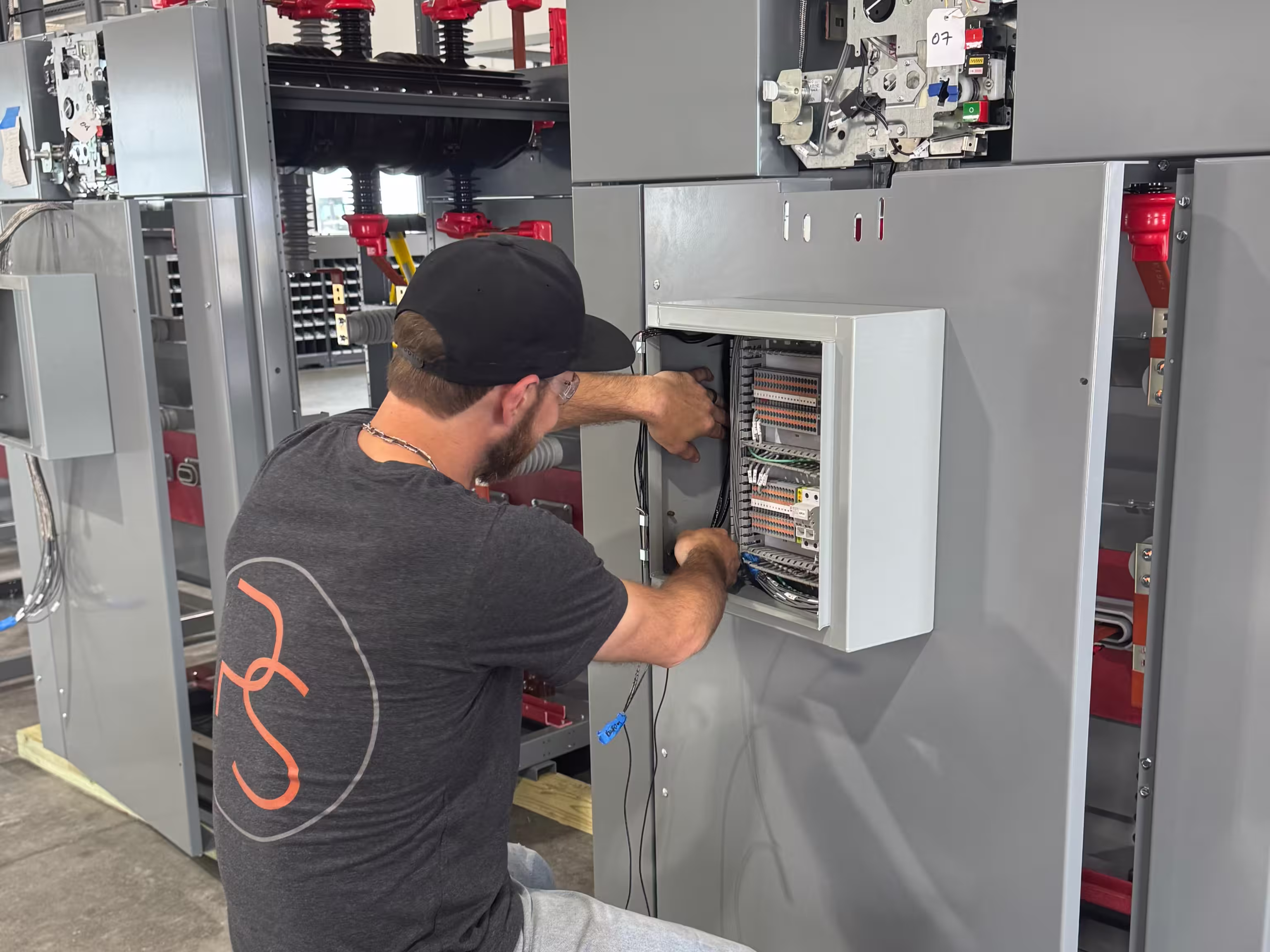 A man wearing a black cap and grey T-shirt with an orange logo is working on wiring inside an open electrical control panel mounted on a large gray industrial machine.