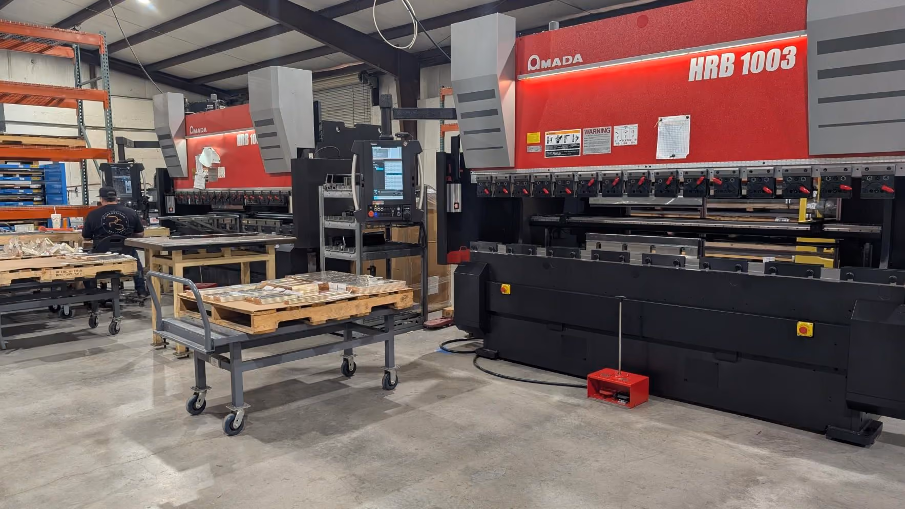 Industrial workspace with two large red Amada HRB 1003 metal press brake machines, a pallet on a wheeled cart, and shelving with materials, on a concrete floor.