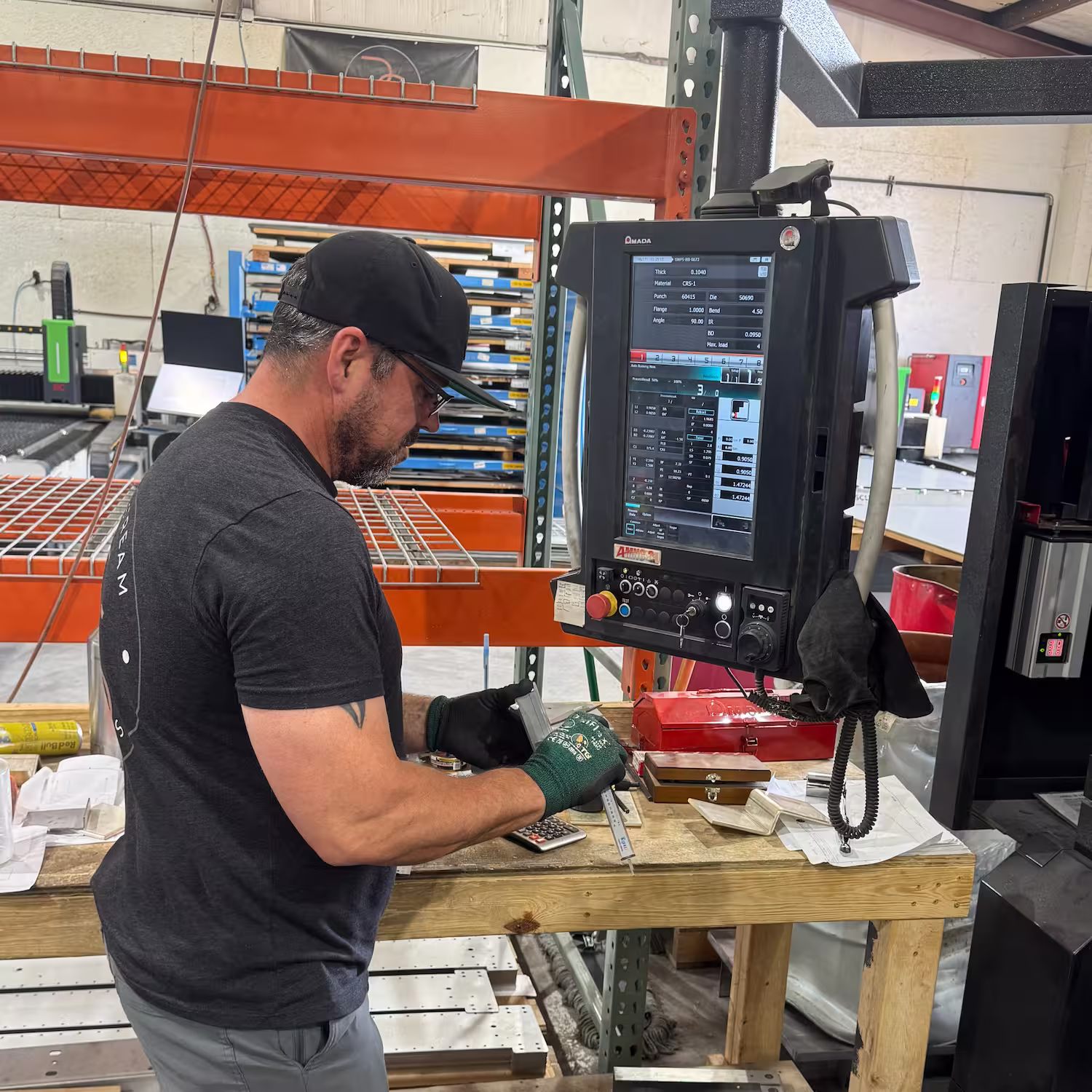 Man wearing a black cap, black t-shirt, and gloves using a caliper to measure an object on a workbench in an industrial workshop with a large machine control panel nearby.