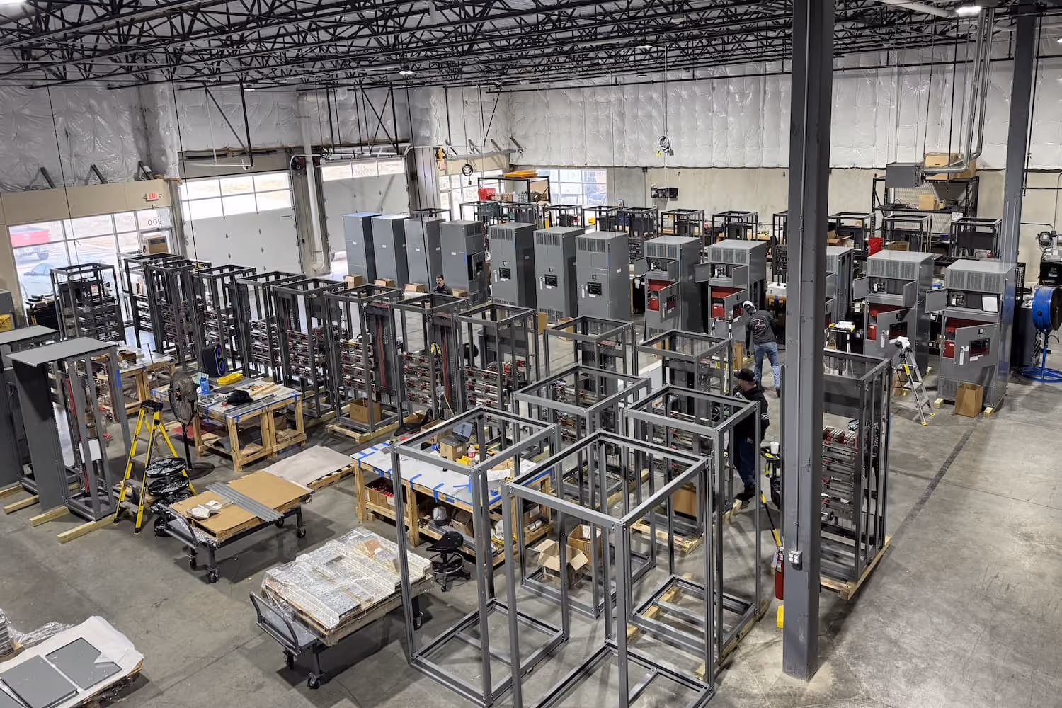 Factory floor with rows of metal cabinet frames and assembled electrical units, workers assembling components amid industrial equipment and tools.